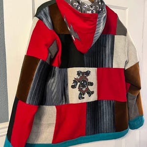 May include: A patchwork hoodie with a variety of fabrics in red, black, brown, gray, and teal. The hood lining features a gray and white pattern. A lightning bolt design is on the hood, and a graphic of a bear is on the back.