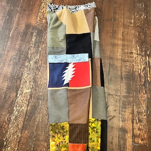 以下が含まれることがあります： 緑、茶色、黒、ベージュの色合いの生地の正方形で構成されたパッチワークスカート。スカートは、花柄、ペイズリー柄、赤と青のグラフィックなど、さまざまなパターンが特徴です。スカートは木製の表面に表示されています。