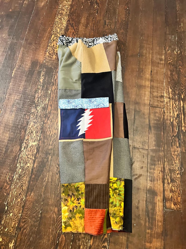 May include: A patchwork skirt composed of various fabric squares in shades of green, brown, black, and tan. The skirt features a mix of patterns, including floral, paisley, and a red and blue graphic. The skirt is displayed on a wooden surface.
