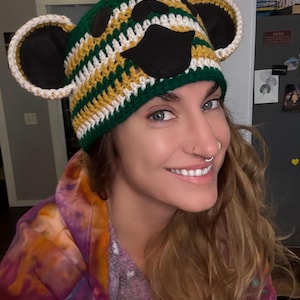 May include: A handmade crochet hat in the shape of a bear's head. The hat features green, yellow, and white stripes, with black felt details for the eyes and nose. The ears are outlined in white.