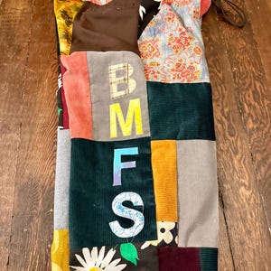 Pu&ograve; includere: Una borsa a coulisse patchwork con una variet&agrave; di tessuti, tra cui velluto a coste e stampe floreali. La borsa presenta le lettere "BMFS" in un carattere in grassetto. La borsa si chiude con un cordoncino.