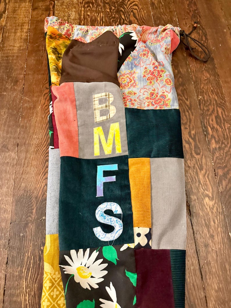 Pu&ograve; includere: Una borsa a coulisse patchwork con una variet&agrave; di tessuti, tra cui velluto a coste e stampe floreali. La borsa presenta le lettere "BMFS" in un carattere in grassetto. La borsa si chiude con un cordoncino.