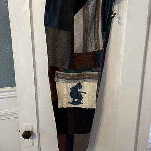 May include: Patchwork pants with a drawstring waist and a large pocket on the left leg. The pocket has a white background with a blue silhouette of a person surfing.