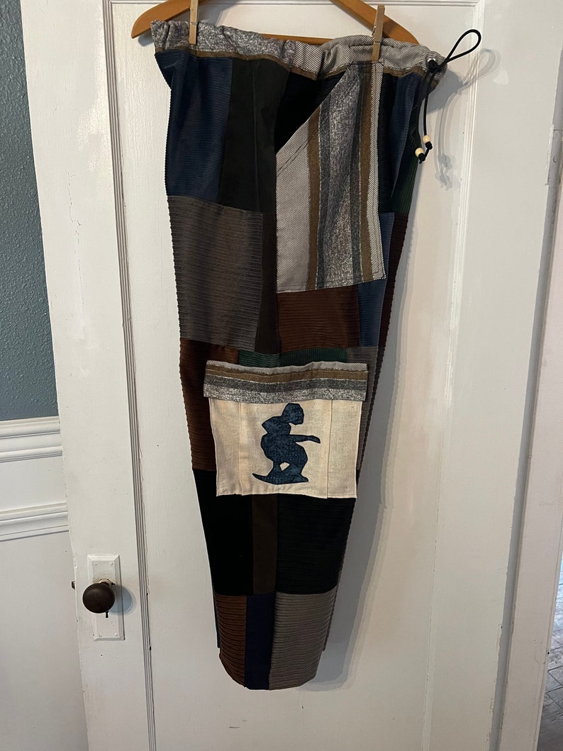 May include: Patchwork pants with a drawstring waist and a large pocket on the left leg. The pocket has a white background with a blue silhouette of a person surfing.