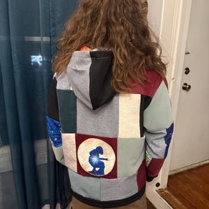 Puede incluir: Una sudadera con capucha de patchwork con una variedad de colores y patrones, incluyendo gris, negro, rojo y azul. La parte trasera de la sudadera con capucha presenta un parche circular con una silueta azul de una persona. La sudadera tiene capucha y cremallera.