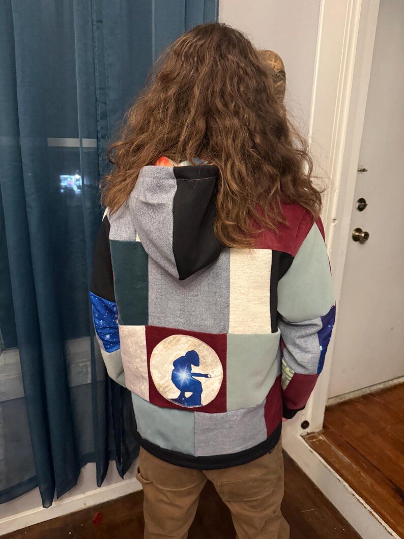 Puede incluir: Una sudadera con capucha de patchwork con una variedad de colores y patrones, incluyendo gris, negro, rojo y azul. La parte trasera de la sudadera con capucha presenta un parche circular con una silueta azul de una persona. La sudadera tiene capucha y cremallera.