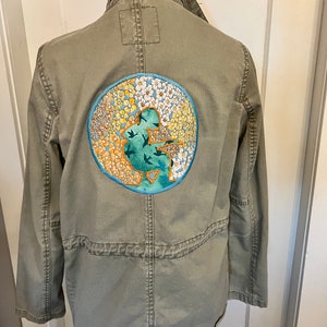 Puede incluir: Chaqueta verde oliva con un parche bordado redondo en la espalda. El parche presenta una figura turquesa con pájaros y un diseño floral en amarillo, blanco y naranja, sobre un fondo azul.