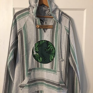 Pode incluir: Camisola de moletom cinza com capuz com um padrão listrado verde e preto. A camisola tem um círculo verde grande com um design de folhas e o texto "WSMFP" em uma etiqueta marrom.