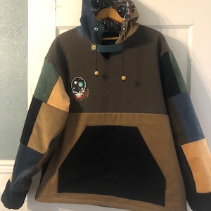Op de afbeelding: Een bruine, groene, blauwe en beige patchwork hoodie met een zwarte zak en een Grateful Dead-logo op de voorkant. De capuchon is gevoerd met een blauwe stof met een zwart-witte print.