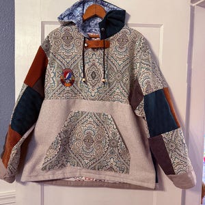 Pode incluir: Um moletom com capuz em patchwork com um grande padrão paisley em tons de azul, verde e marrom. O moletom tem um grande bolso na frente e um pequeno patch com o logotipo do Grateful Dead no peito esquerdo.