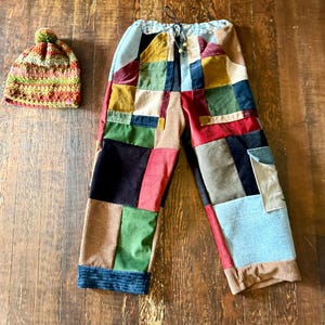 May include: A pair of patchwork pants with a drawstring waist and a matching crocheted beanie. The pants feature a variety of colorful corduroy and velvet patches. The beanie is a mix of green, orange, and brown yarn with a pom-pom.