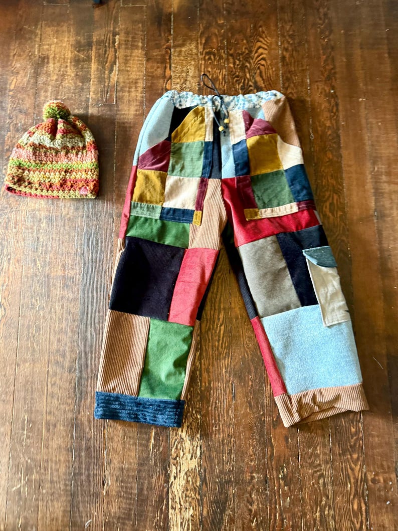 May include: A pair of patchwork pants with a drawstring waist and a matching crocheted beanie. The pants feature a variety of colorful corduroy and velvet patches. The beanie is a mix of green, orange, and brown yarn with a pom-pom.