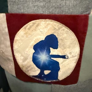 Puede incluir: Un dise&ntilde;o de patchwork con un parche circular con una silueta azul de una persona tocando un instrumento contra un fondo estrellado. El parche est&aacute; sobre un fondo rojo, con tela gris, verde y blanca.