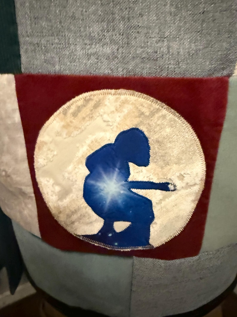 Puede incluir: Un dise&ntilde;o de patchwork con un parche circular con una silueta azul de una persona tocando un instrumento contra un fondo estrellado. El parche est&aacute; sobre un fondo rojo, con tela gris, verde y blanca.