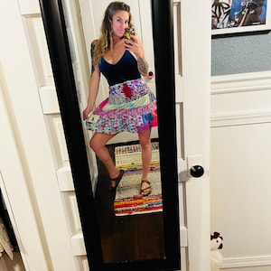 May include: A woman wearing a blue velvet tank top and a colorful patchwork skirt. She is standing in front of a mirror and is wearing sandals.