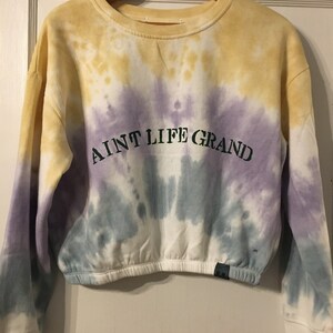 Puede incluir: Una sudadera teñida con tintes amarillos, morados y azules. La sudadera corta presenta el texto "AINT LIFE GRAND" en letras verdes. La parte superior de manga larga está colgada en una percha de madera.