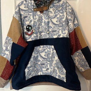 May include: A blue, brown, and white patchwork hoodie with a paisley pattern. The hoodie has a large pocket on the front and a drawstring hood. The hood is lined with a black and white fabric with a skull and crossbones design.