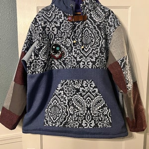 May include: A blue and gray patchwork hoodie with a paisley print and a small embroidered patch. The hoodie has a kangaroo pocket and a drawstring hood.