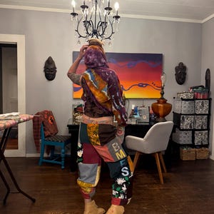 Pu&ograve; includere: Una persona indossa un top con cappuccio tie-dye e pantaloni patchwork. I pantaloni presentano vari quadrati di tessuto nelle tonalit&agrave; del marrone, rosso e verde, con dettagli floreali e di lettere. Stivali color cammello completano l'outfit. Un lampadario &egrave; appeso sopra.