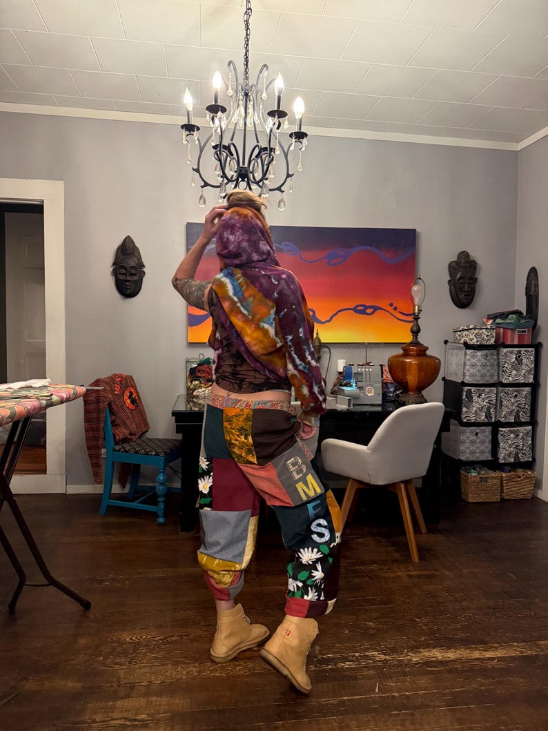 Pu&ograve; includere: Una persona indossa un top con cappuccio tie-dye e pantaloni patchwork. I pantaloni presentano vari quadrati di tessuto nelle tonalit&agrave; del marrone, rosso e verde, con dettagli floreali e di lettere. Stivali color cammello completano l'outfit. Un lampadario &egrave; appeso sopra.