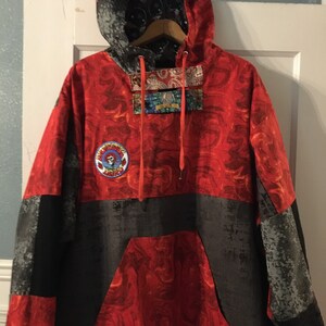 May include: A red, black, and gray hooded sweatshirt with a patchwork design. The sweatshirt has a large pocket on the front and a small patch with a cartoon bear on the left side. The hood is lined with a black and white patterned fabric.