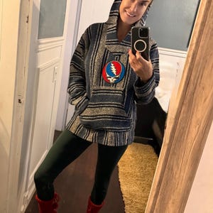 Peut inclure: Un sweat à capuche rayé bleu et gris avec une poche avant et une capuche. Le sweat à capuche présente un écusson coloré avec un éclair et un crâne. La personne porte des leggings vert foncé et des bottes rouges.