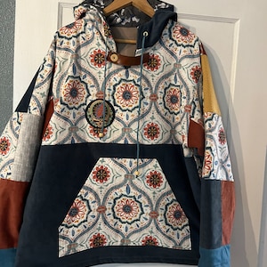 May include: A blue and white patterned hoodie with a large pocket on the front. The hoodie has a brown leather strap and a drawstring. The fabric is a patchwork of different patterns.