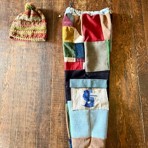 May include: A colorful patchwork pair of pants with a drawstring waist and a matching crocheted beanie. The pants feature various fabric patches in shades of red, blue, green, and brown. The beanie is a mix of green, red, and brown yarn.