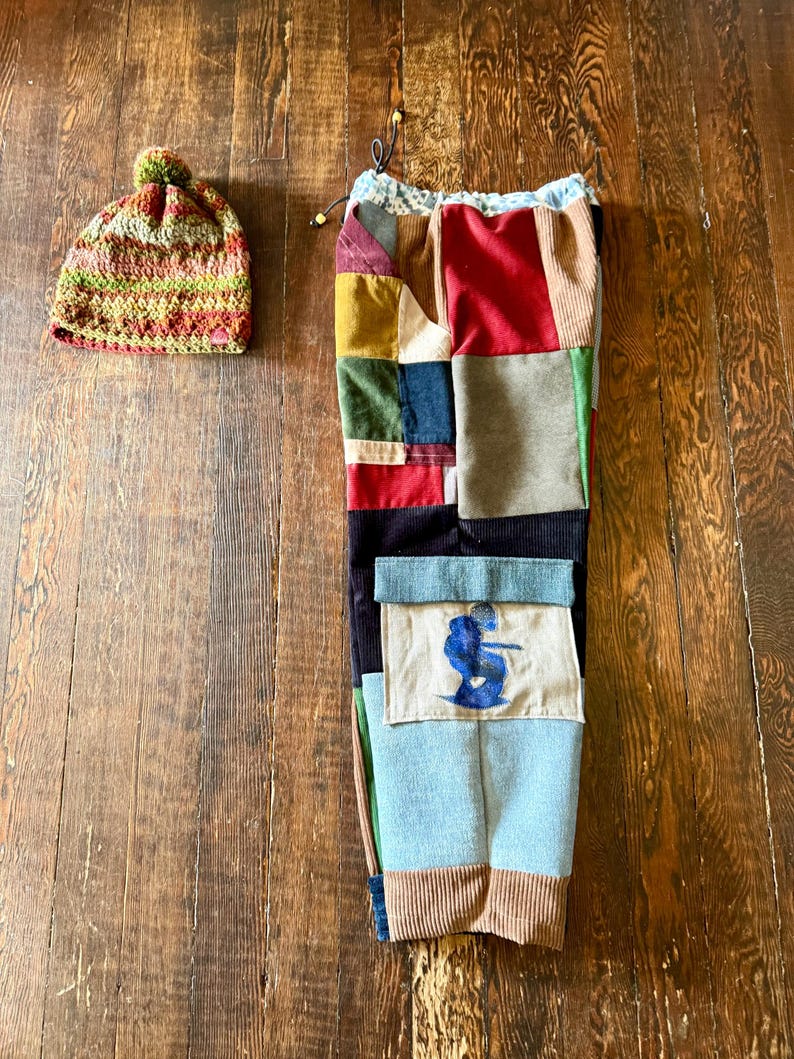 May include: A colorful patchwork pair of pants with a drawstring waist and a matching crocheted beanie. The pants feature various fabric patches in shades of red, blue, green, and brown. The beanie is a mix of green, red, and brown yarn.