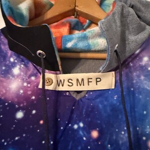 Puede incluir: Una sudadera con capucha con un estampado de galaxia en tonos azules, morados y blancos. El forro de la capucha presenta una tela estampada colorida en naranja, azul y blanco. La etiqueta dice "WSMFP".
