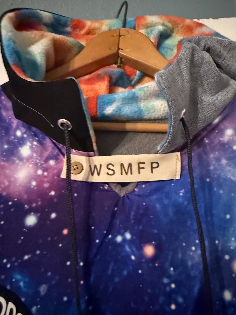 Puede incluir: Una sudadera con capucha con un estampado de galaxia en tonos azules, morados y blancos. El forro de la capucha presenta una tela estampada colorida en naranja, azul y blanco. La etiqueta dice "WSMFP".