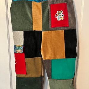 May include: Patchwork corduroy pants with a variety of colors and patterns, including green, brown, black, red, and blue. A small red pocket with a tie-dye bear patch is on the right side of the pants.