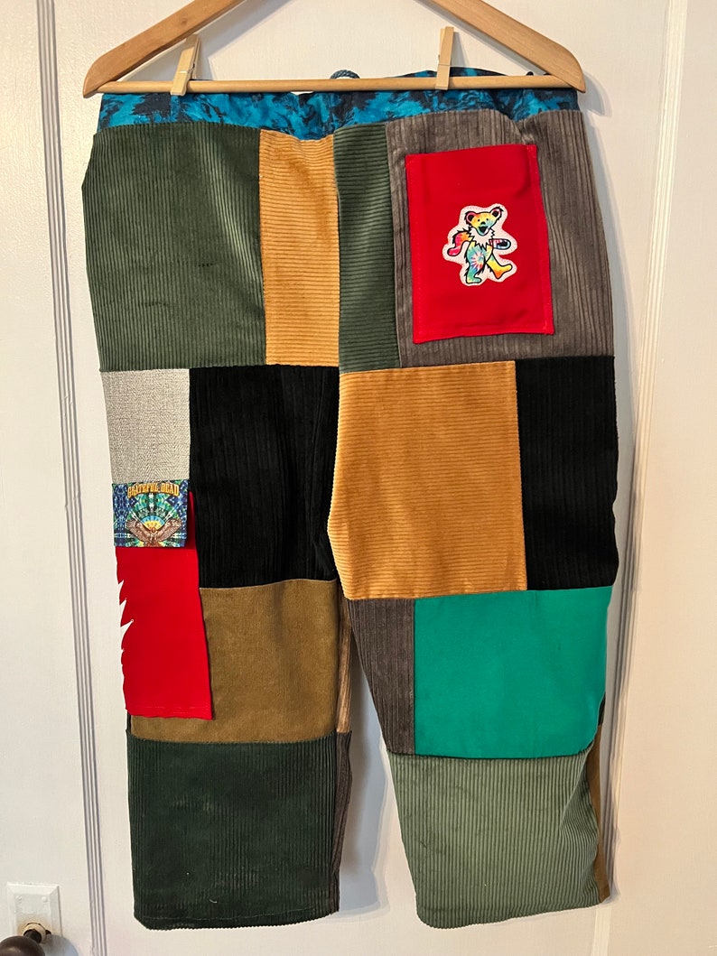 May include: Patchwork corduroy pants with a variety of colors and patterns, including green, brown, black, red, and blue. A small red pocket with a tie-dye bear patch is on the right side of the pants.