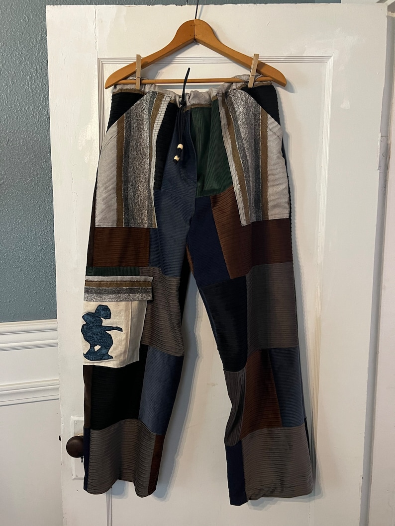 May include: Patchwork pants with a drawstring waist and a variety of colors and textures, including brown, blue, green, and white. The pants have a small embroidered design on the left leg.