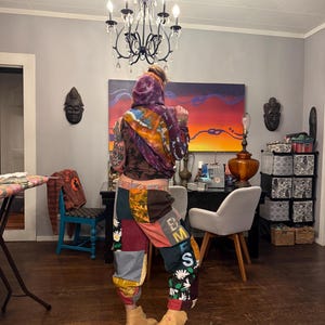 Pu&ograve; includere: Una persona indossa una felpa con cappuccio tie-dye e pantaloni patchwork con le lettere "EMS" sul lato. I pantaloni sono un mix di colori e fantasie, con accenti floreali. Stivali color cammello completano il look.