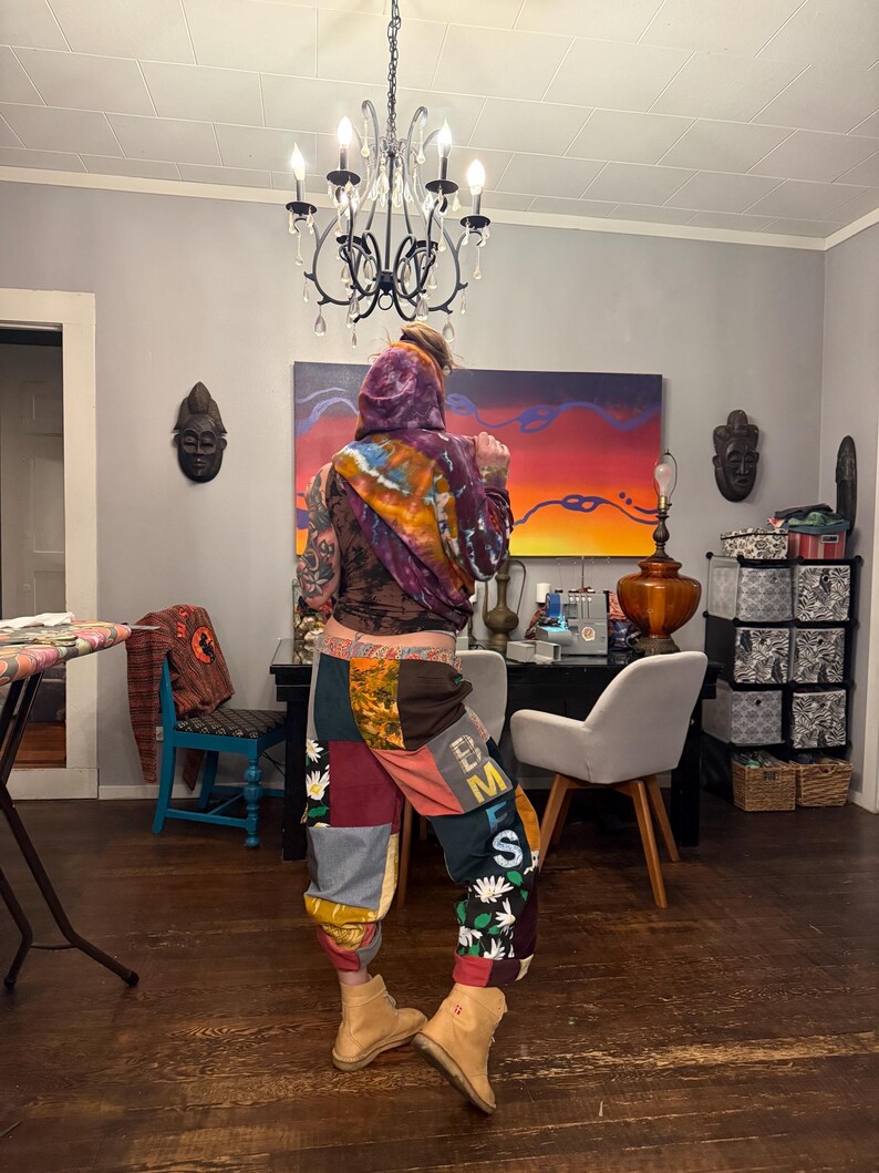 Pu&ograve; includere: Una persona indossa una felpa con cappuccio tie-dye e pantaloni patchwork con le lettere "EMS" sul lato. I pantaloni sono un mix di colori e fantasie, con accenti floreali. Stivali color cammello completano il look.