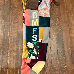 Peut inclure: Vêtement patchwork avec une variété de tissus et de couleurs. Le vêtement présente les lettres "BMFS" disposées verticalement. Les tissus comprennent des imprimés floraux, du velours côtelé et des couleurs unies comme le rose, le vert et le jaune. Un cordon de serrage est visible en haut.