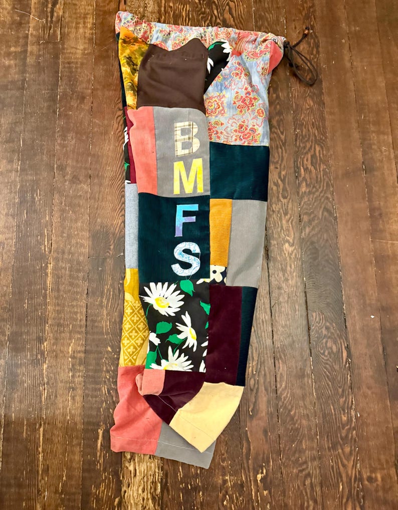 Pu&ograve; includere: Un capo patchwork con una variet&agrave; di tessuti e colori. Il capo presenta le lettere "BMFS" in disposizione verticale. I tessuti includono stampe floreali, velluto a coste e colori uniti come rosa, verde e giallo. Un cordoncino &egrave; visibile nella parte superiore.