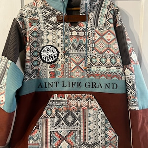 Könnte beinhalten: Ein mehrfarbiger Patchwork-Hoodie mit einer großen Tasche auf der Vorderseite. Der Hoodie hat eine blaue Kapuze und ein braunes Patch mit dem Text "WGMNTS" auf der Rückseite des Halses. Die Vorderseite des Hoodies hat ein schwarzes Patch mit dem Text "AUDESPREAD PATPJIC" und den Text "AIN'T LIFE GRAND" in Schwarz auf einem blauen Band.