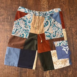 May include: Patchwork shorts featuring a variety of fabrics including blue paisley, solid brown, dark green, and navy corduroy. The shorts have an elastic waistband with a green drawstring and a blue pocket patch displaying a white goat head logo.
