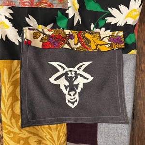 Pu&ograve; includere: Tessuto patchwork con una tasca grigia con un disegno a testa di capra bianca. Il patchwork include tessuti floreali e fantasia in vari colori, tra cui nero, giallo, rosso e grigio. La tasca &egrave; cucita sul patchwork.