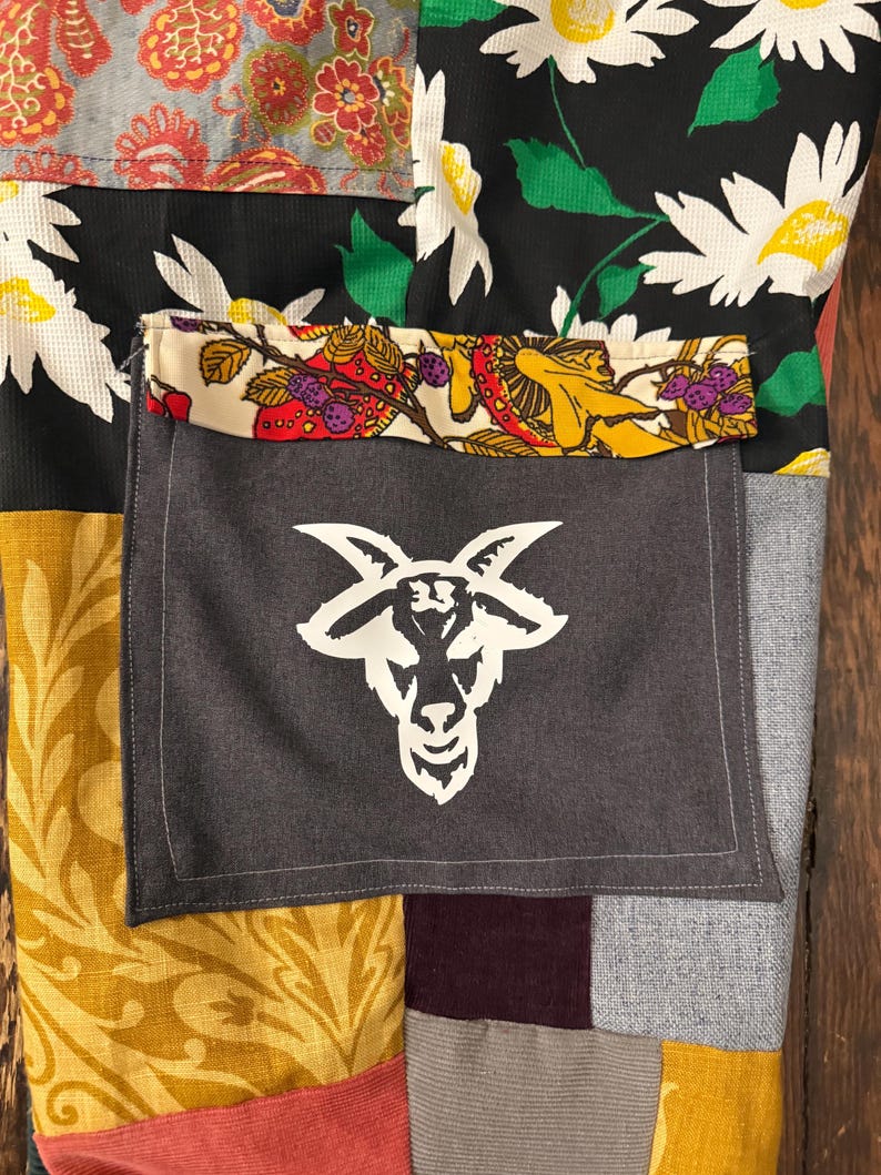 Pu&ograve; includere: Tessuto patchwork con una tasca grigia con un disegno a testa di capra bianca. Il patchwork include tessuti floreali e fantasia in vari colori, tra cui nero, giallo, rosso e grigio. La tasca &egrave; cucita sul patchwork.