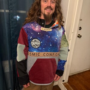 Puede incluir: Una sudadera con capucha multicolor con un estampado de galaxia en el pecho y la capucha. La sudadera con capucha presenta un gran bolsillo delantero y parches con el texto "Widespread Panic" y "Cosmic Confidant". Las mangas son una mezcla de colores, incluyendo gris, burdeos y verde.