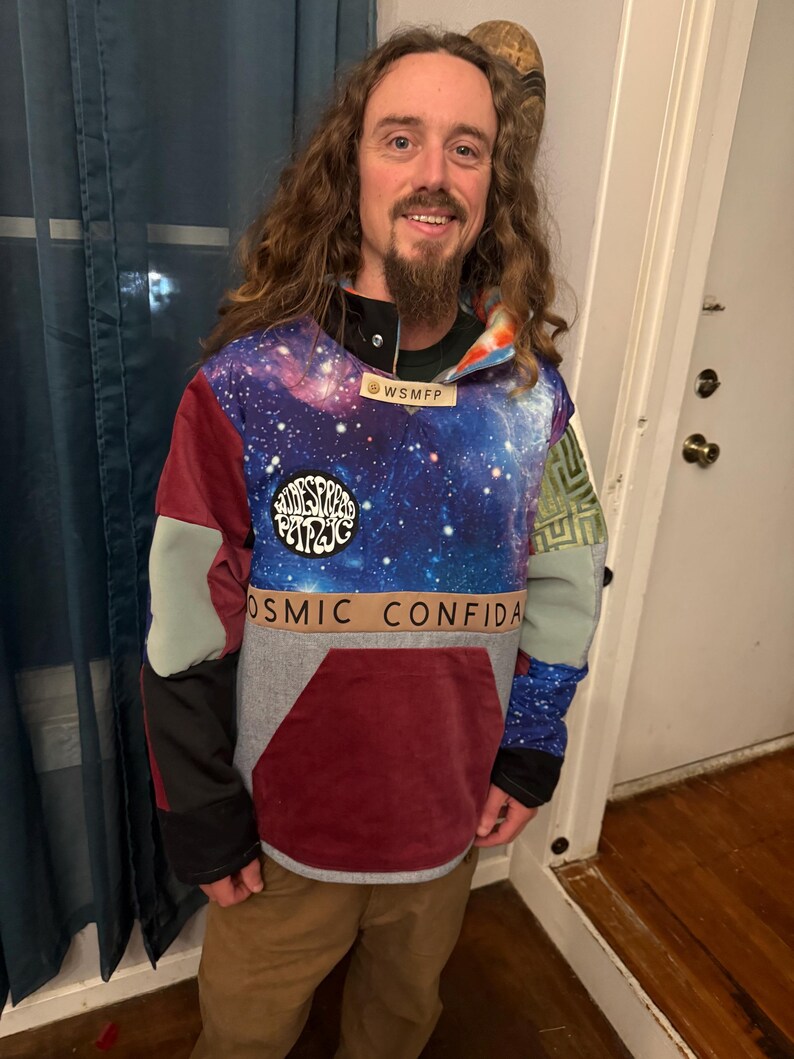 Puede incluir: Una sudadera con capucha multicolor con un estampado de galaxia en el pecho y la capucha. La sudadera con capucha presenta un gran bolsillo delantero y parches con el texto "Widespread Panic" y "Cosmic Confidant". Las mangas son una mezcla de colores, incluyendo gris, burdeos y verde.