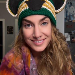 May include: A handmade crochet hat in green, yellow, white, and black, featuring bear ears. The hat has a rounded crown and a white trim around the ears. The hat is designed to be worn on the head.