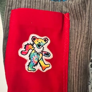 May include: A red fabric patch with a tie-dye bear graphic. The bear is walking and has a smiling face. The patch is sewn onto a gray corduroy fabric.