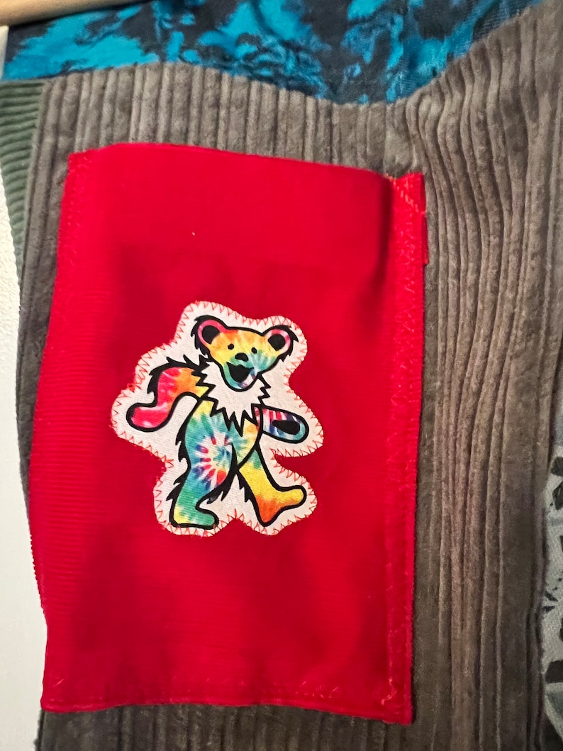 May include: A red fabric patch with a tie-dye bear graphic. The bear is walking and has a smiling face. The patch is sewn onto a gray corduroy fabric.