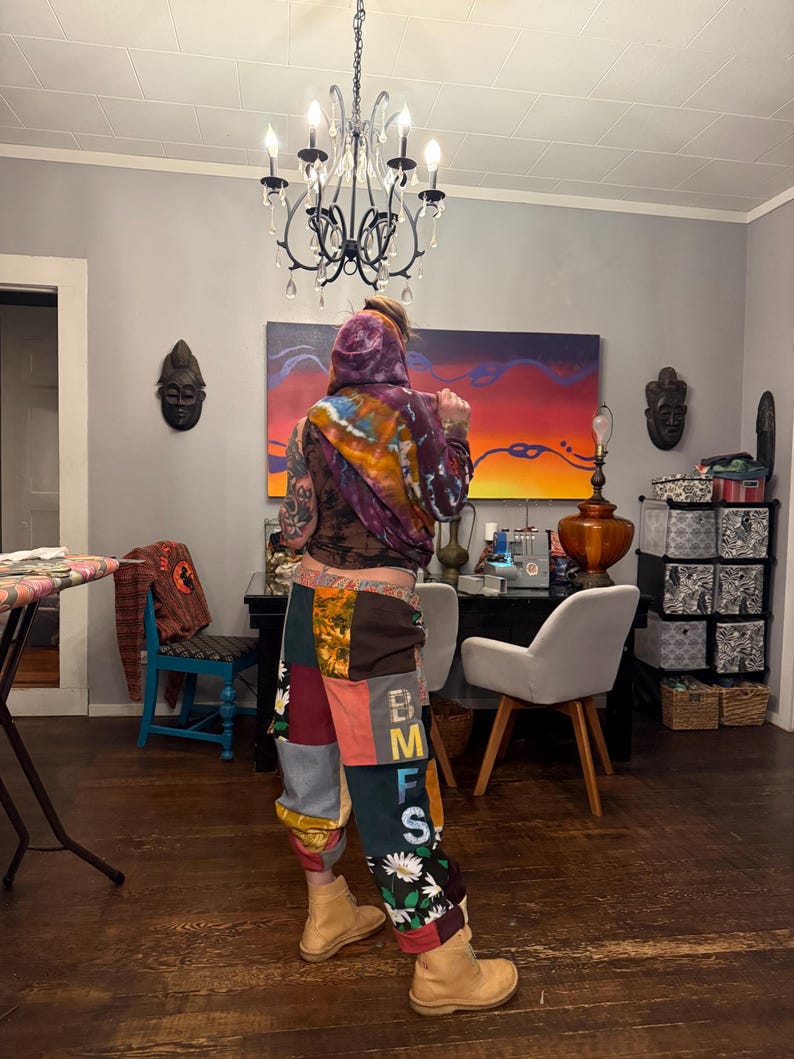 Pu&ograve; includere: Una persona indossa una felpa con cappuccio tie-dye e pantaloni patchwork con le lettere "BMFS" sulla gamba. I pantaloni presentano vari modelli di tessuto, tra cui disegni floreali e geometrici. La persona indossa stivali color cachi.