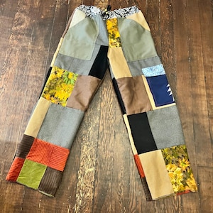 May include: Patchwork pants with a variety of fabrics in shades of green, brown, black, and yellow. The pants have a drawstring waist and a mix of textures, including corduroy and floral prints. The design is a unique, handcrafted style.