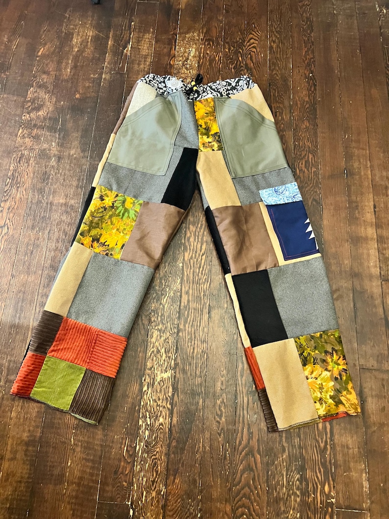 May include: Patchwork pants with a variety of fabrics in shades of green, brown, black, and yellow. The pants have a drawstring waist and a mix of textures, including corduroy and floral prints. The design is a unique, handcrafted style.